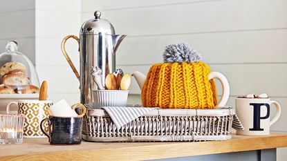 Wooden kitchen worktop with wicker tray holding a teapot in a yellow knitted cover and a chrome coffee jug