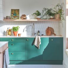 White tiled kitchen with teal painted cabinets underneath a sink and floating shelf