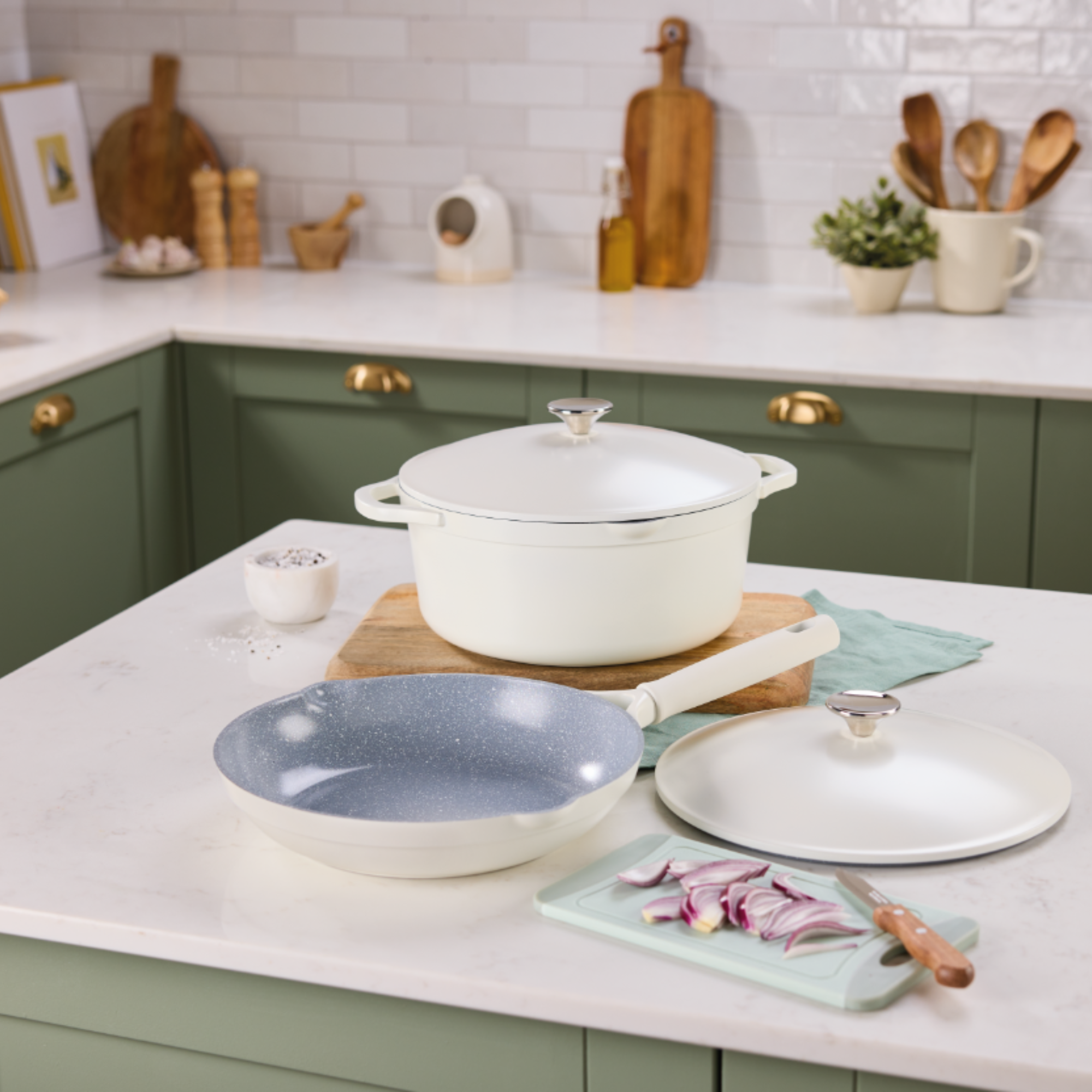 Aldi aluminium frying pan and casserole on a white kitchen island in a kitchen with white countertops and green cupboards.
