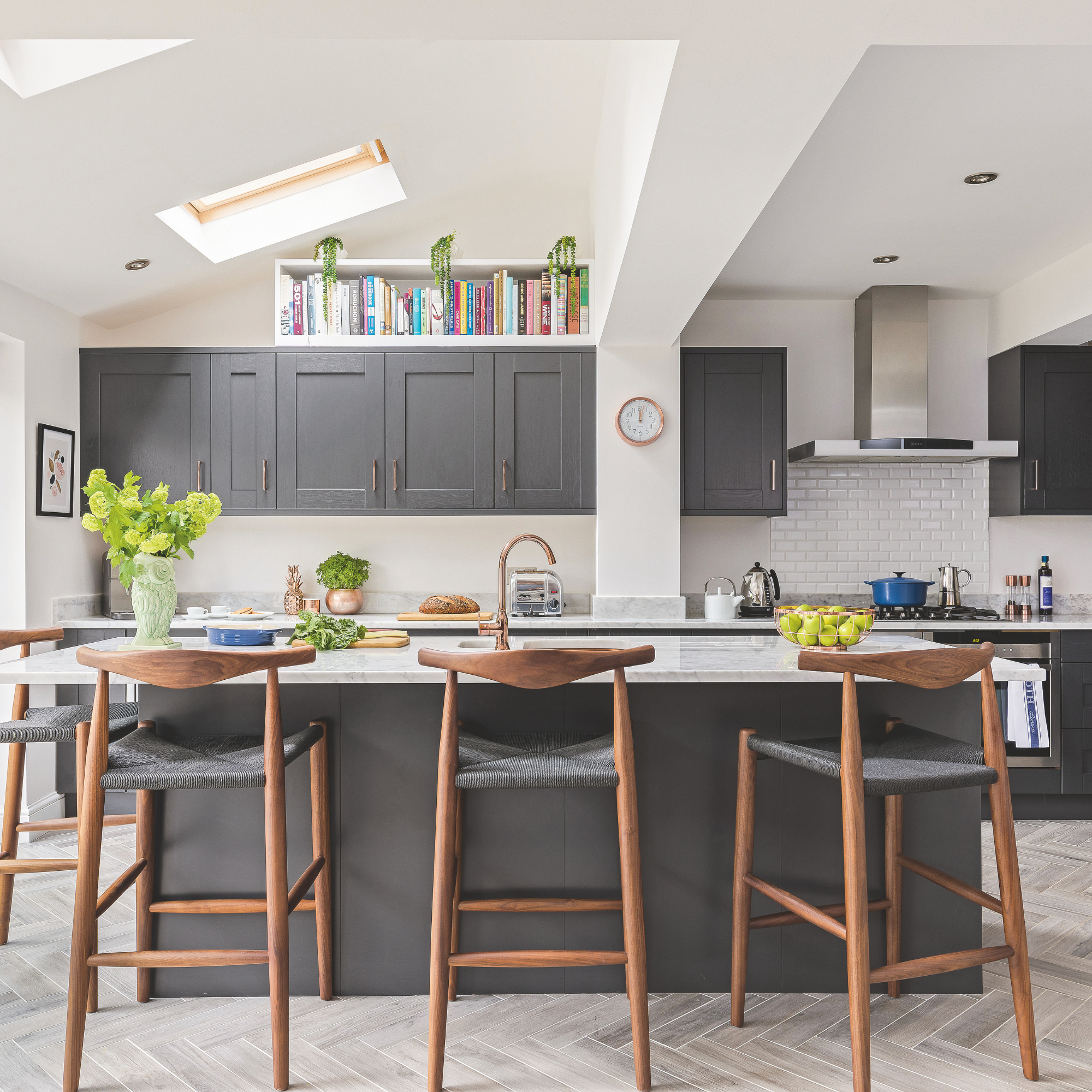 Kitchen with kitchen island, dark grey kitchen cupboards with a white worktop, bar stools in grey and copper and pale grey flooring. Pub Orig A new kitchen in Gemma and Brad Isaac's five bedroom detached 1940's house in Raynes Park, London.